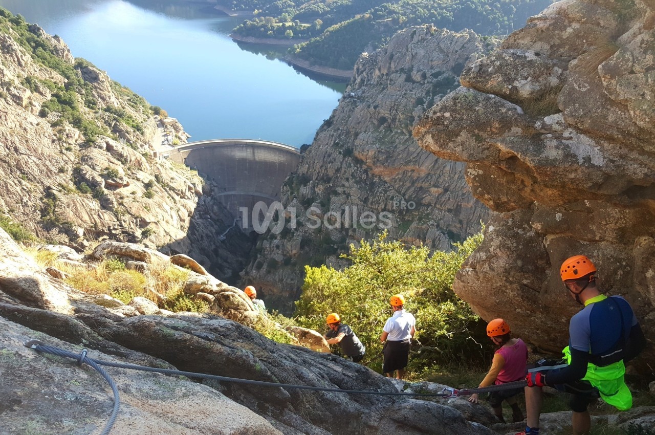 Des personnes équipées pour la descente en rappel dans un paysage montagneux avec un barrage visible au loin.