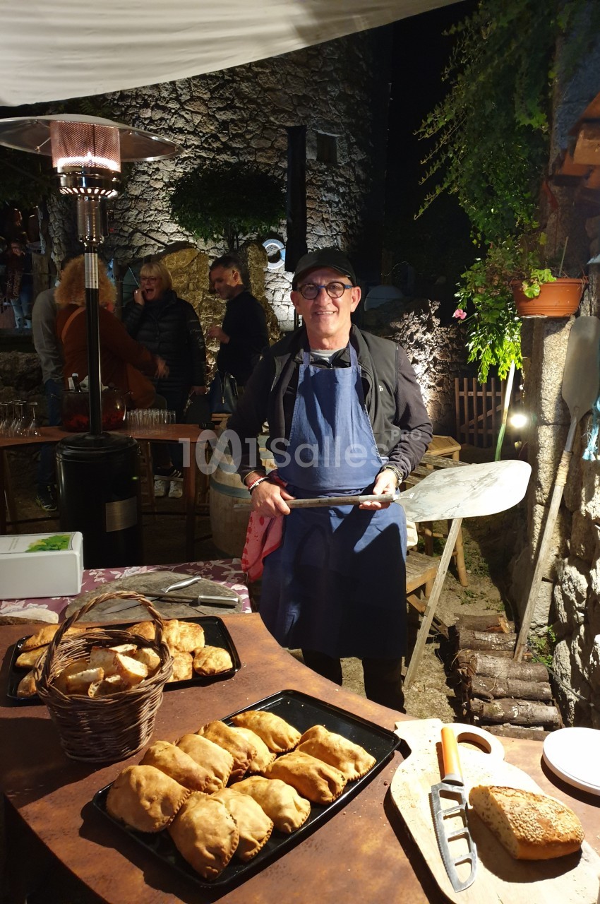 Un homme en tablier sert des plats cuisinés lors d'un événement en plein air, éclairé par une lampe chauffante.