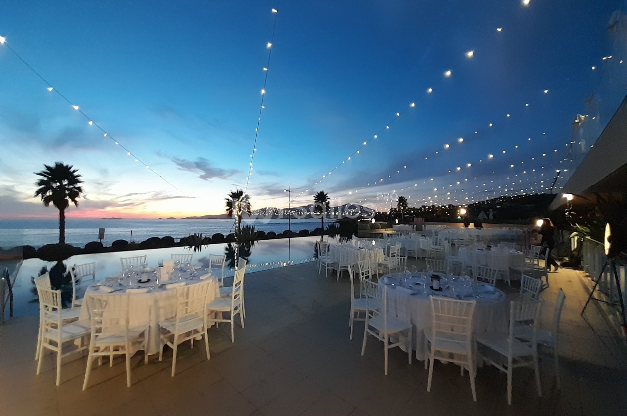Tables dressées en extérieur près d'une piscine, sous des guirlandes lumineuses, au coucher du soleil.