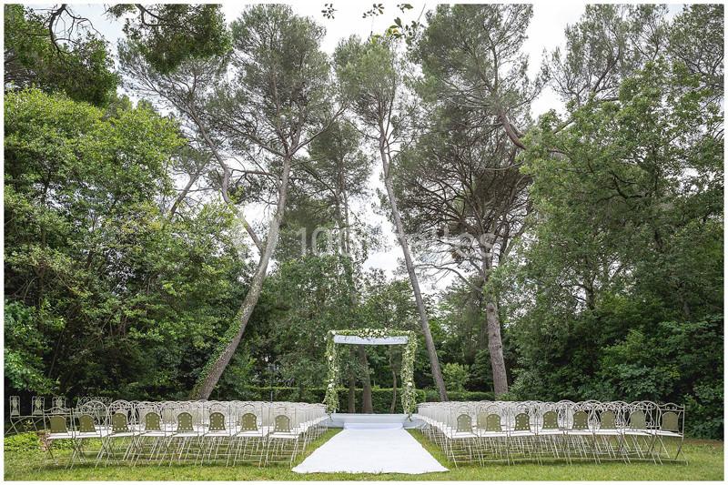 Chaises blanches disposées en rangées face à une arche décorée, dans un cadre naturel entouré d'arbres.