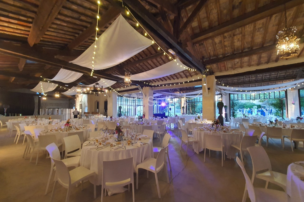 Salle de réception décorée avec des guirlandes lumineuses, des tables rondes dressées et des chaises blanches.