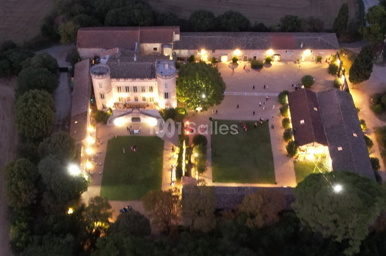 Vue aérienne d'un château éclairé de nuit, entouré de jardins, d'arbres et de bâtiments annexes.