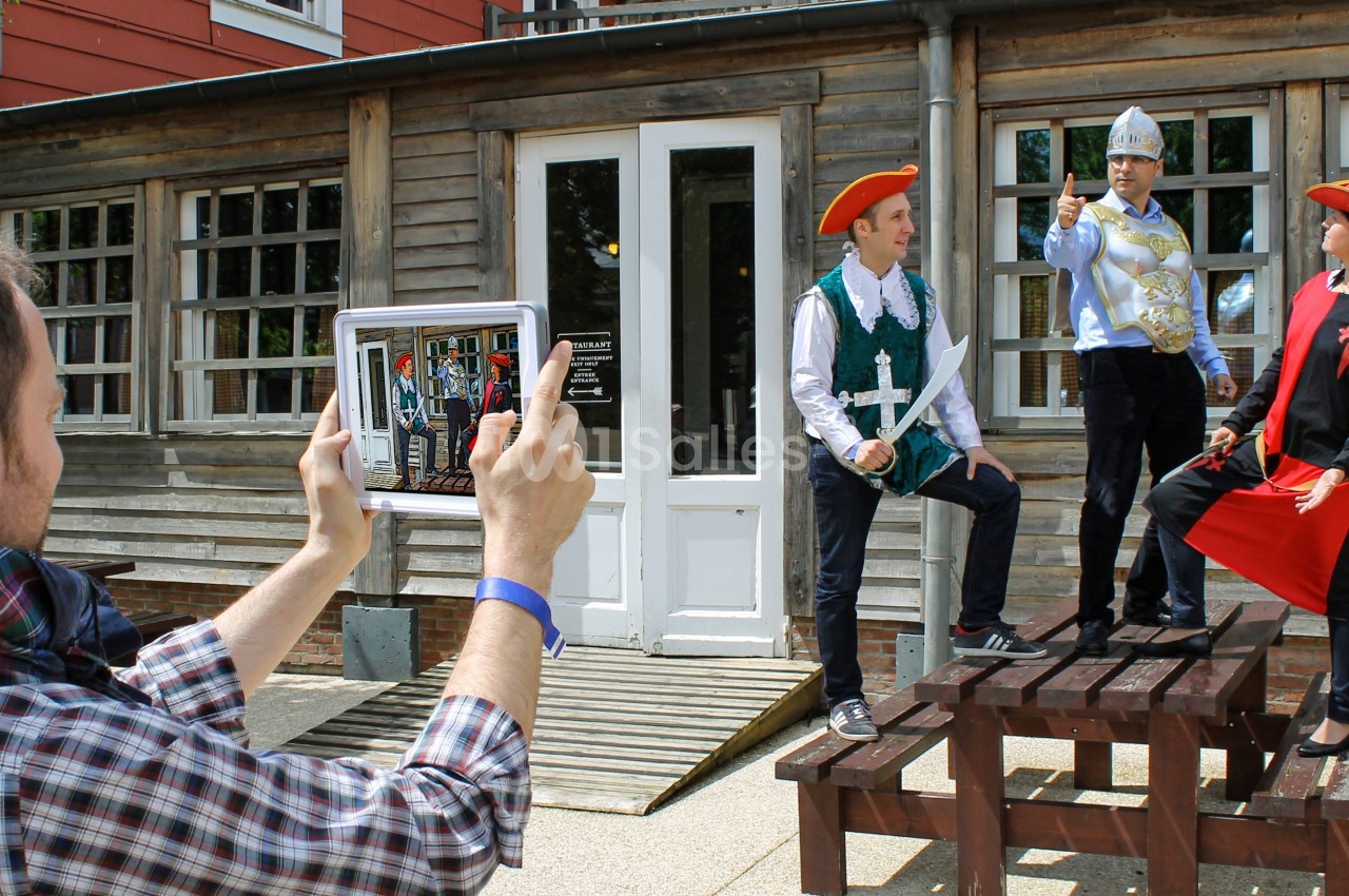 Un homme photographie trois personnes déguisées en costumes historiques posant sur des bancs devant un bâtiment en bois.