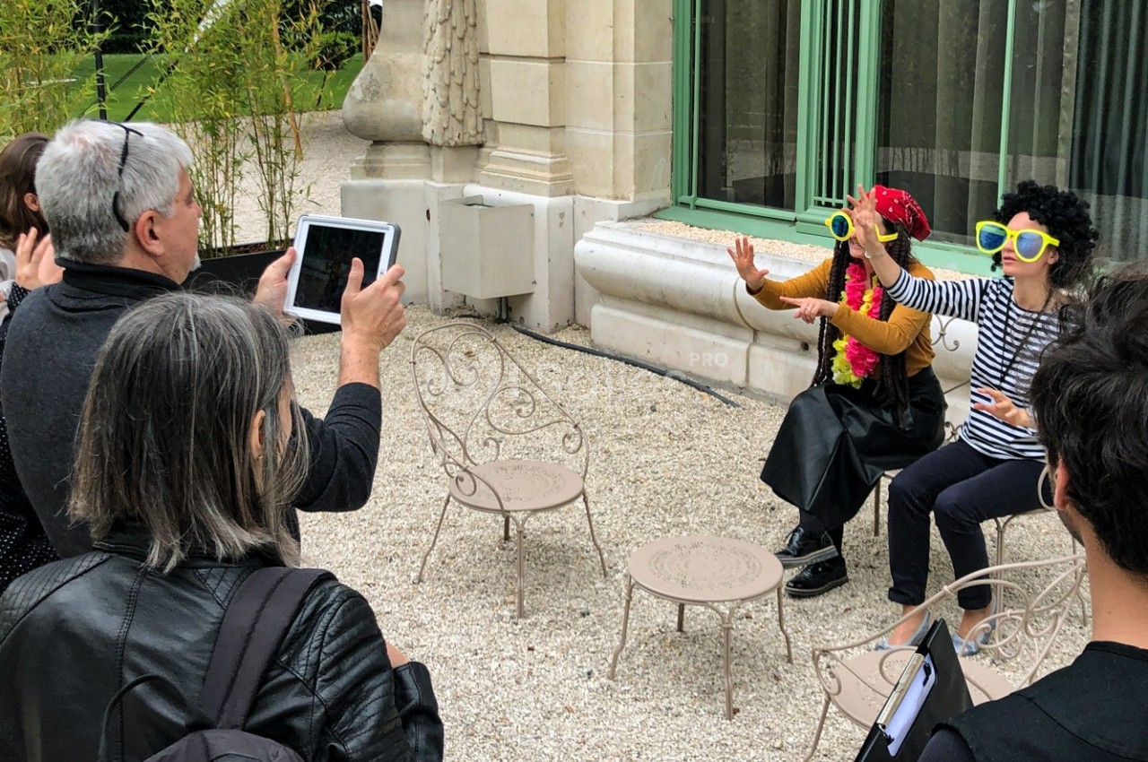 Des personnes photographient deux individus déguisés avec des lunettes colorées, assis sur des chaises en extérieur.