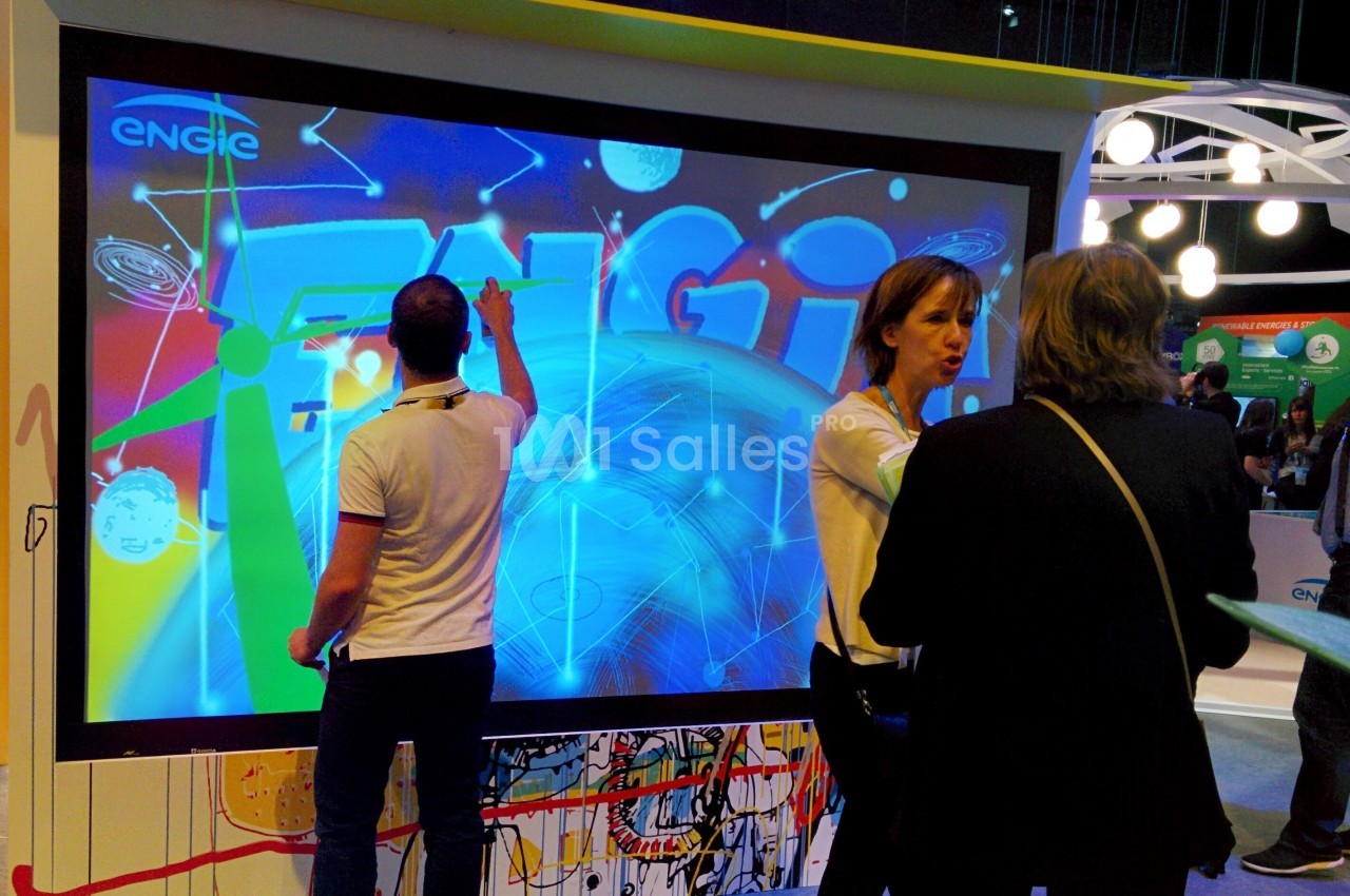 Trois personnes interagissent devant un écran tactile affichant des visuels colorés dans un espace d'exposition.