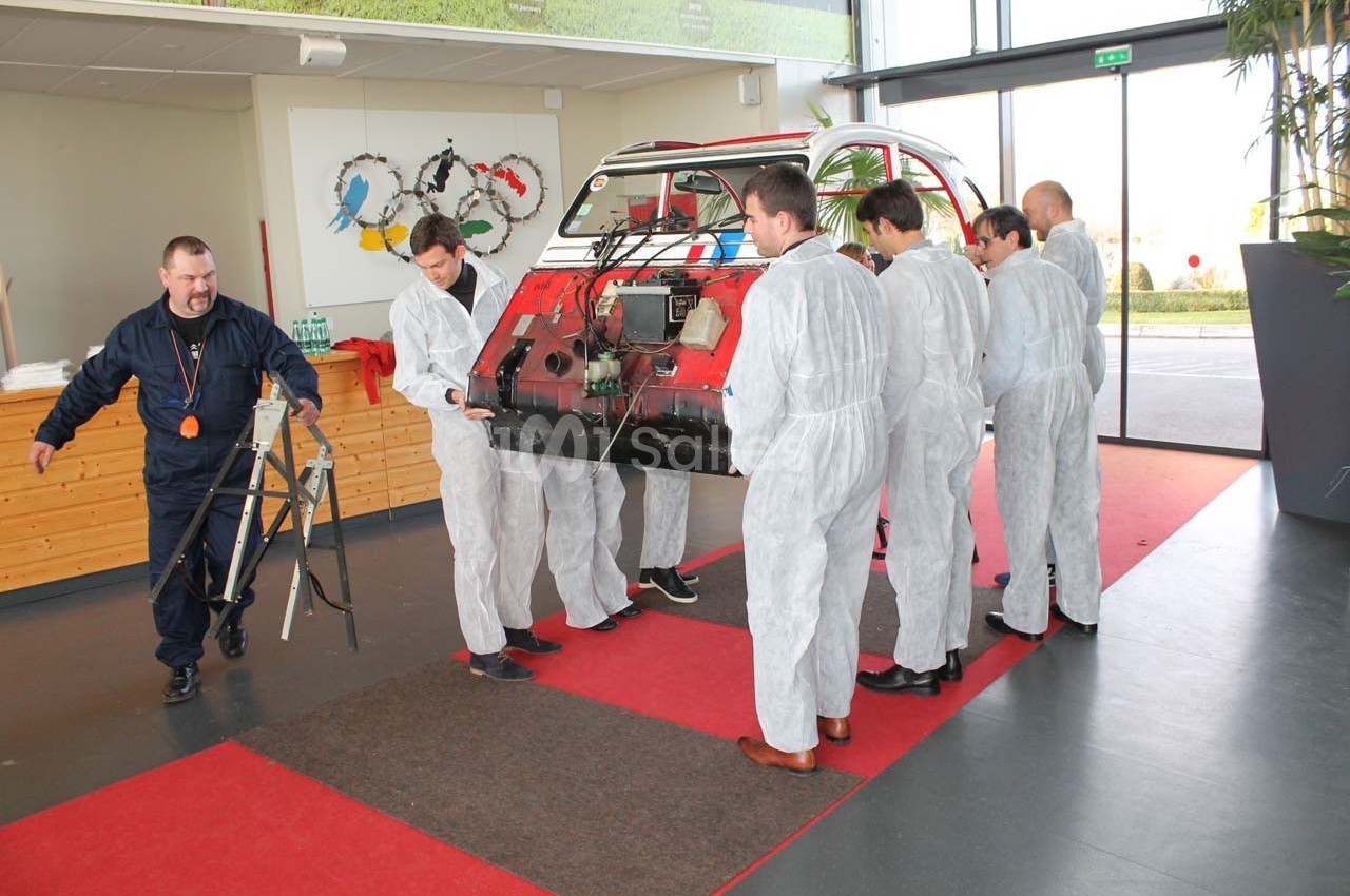 Des personnes en combinaison blanche manipulent une grande pièce de carrosserie rouge dans un espace intérieur lumineux.