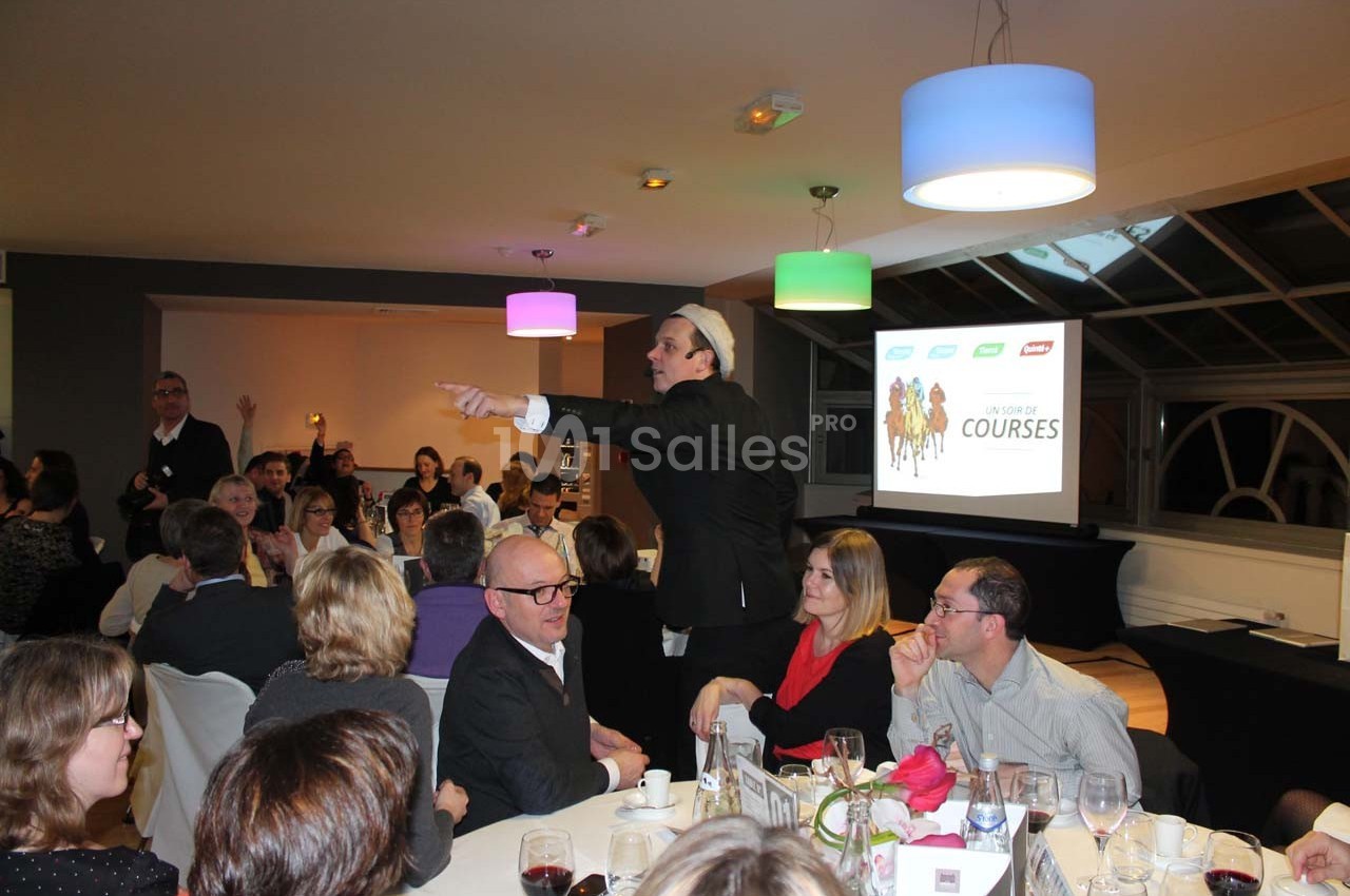 Un homme anime une soirée conviviale avec des participants assis autour de tables dans une salle éclairée.