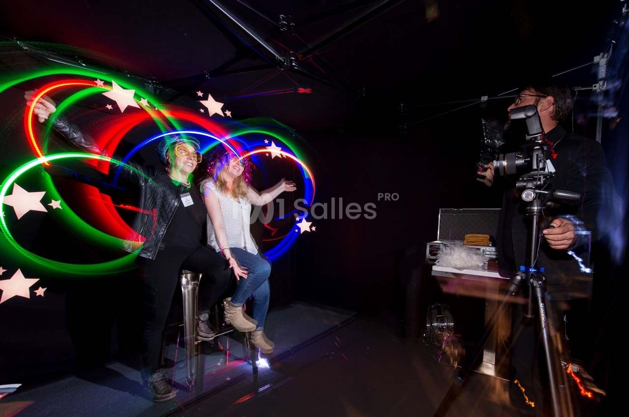 Deux personnes posent sous des lumières colorées en mouvement, avec un photographe équipé d'un appareil sur trépied.