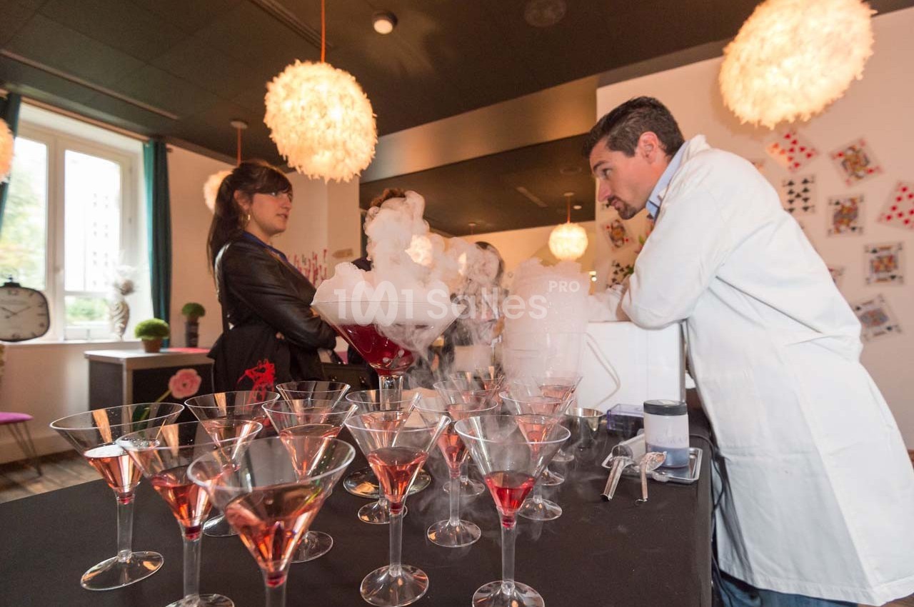 Un homme en blouse blanche prépare des cocktails avec de la fumée devant deux personnes dans un cadre intérieur.