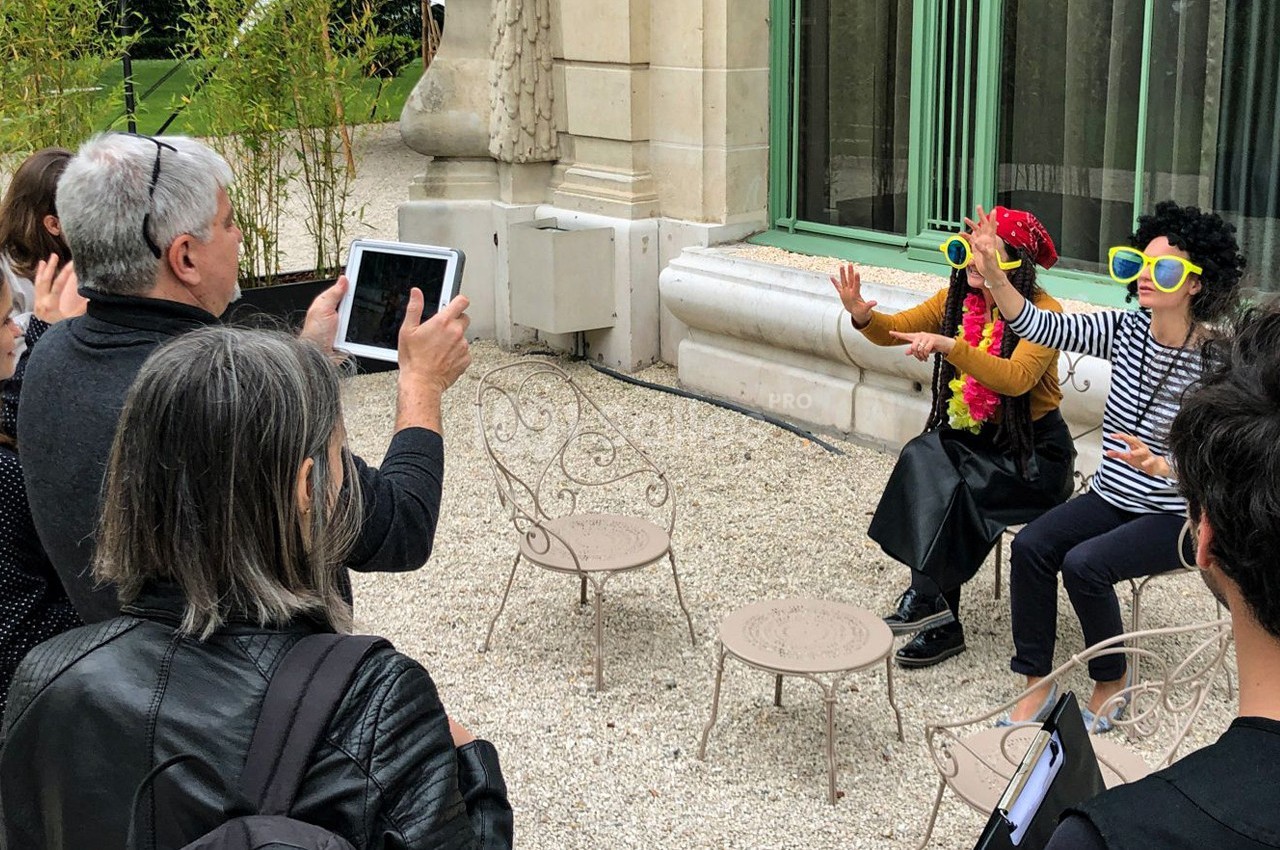 Des personnes photographient deux individus déguisés avec des lunettes colorées, assis sur des chaises en extérieur.
