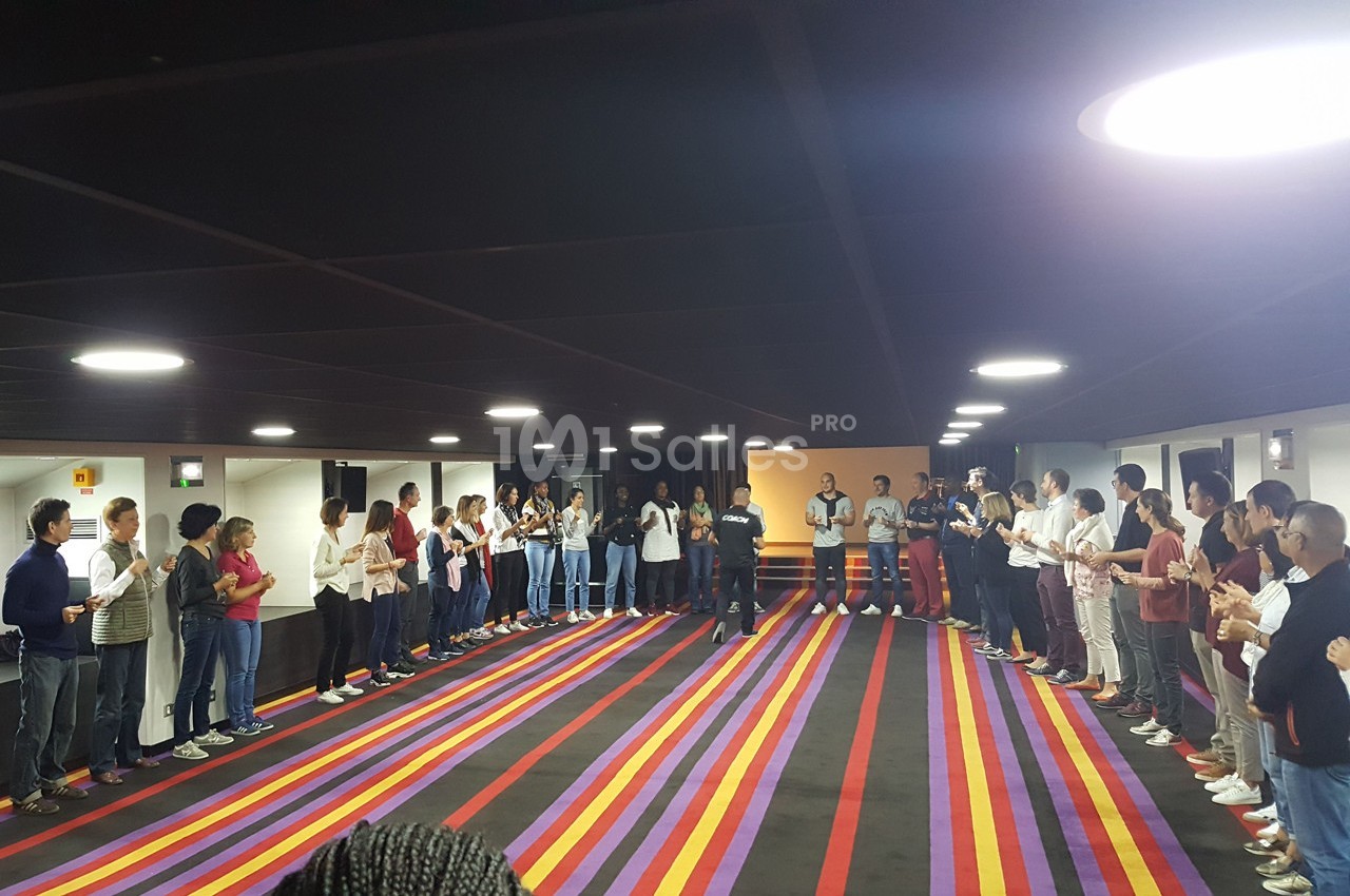 Des personnes debout en cercle dans une grande salle colorée, participant à une activité collective.