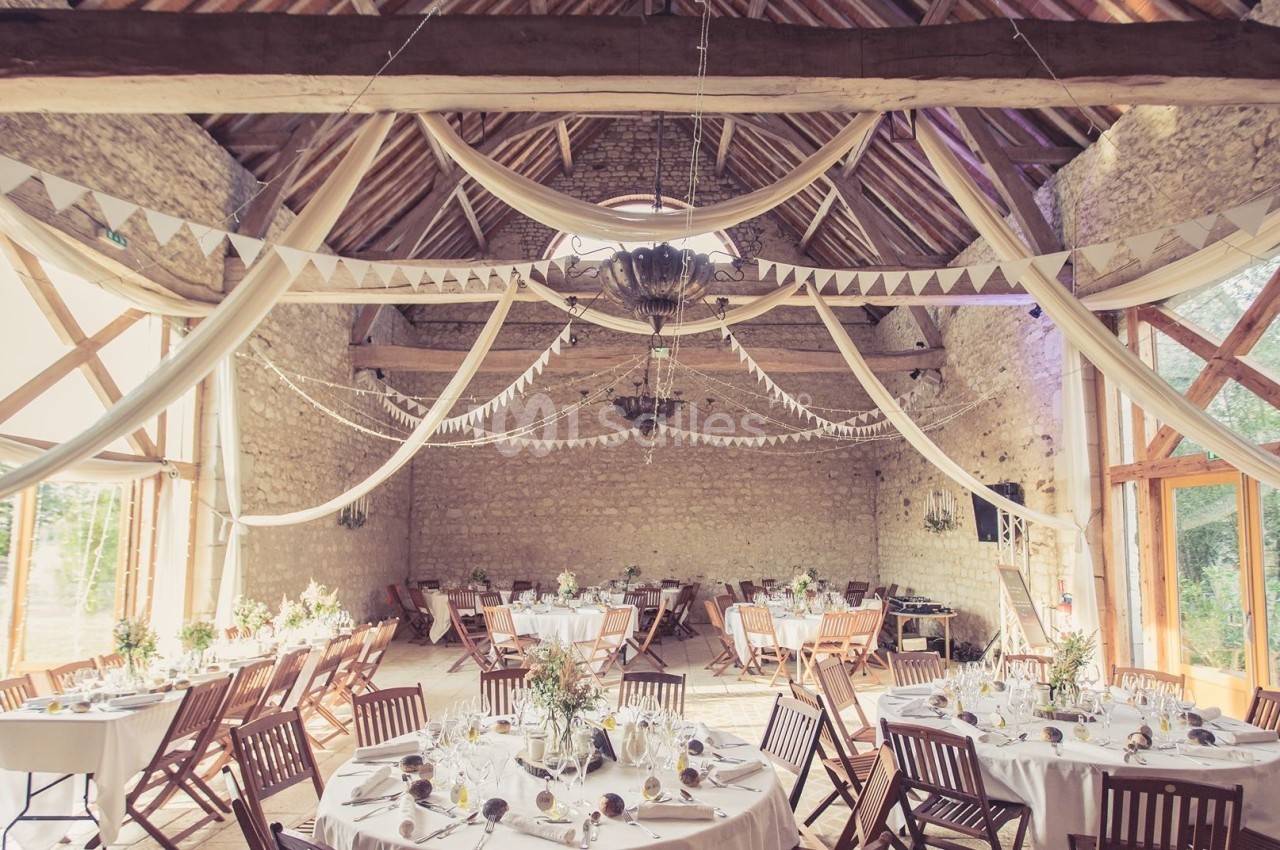 Salle de réception en pierre avec poutres apparentes, tables décorées et guirlandes suspendues.