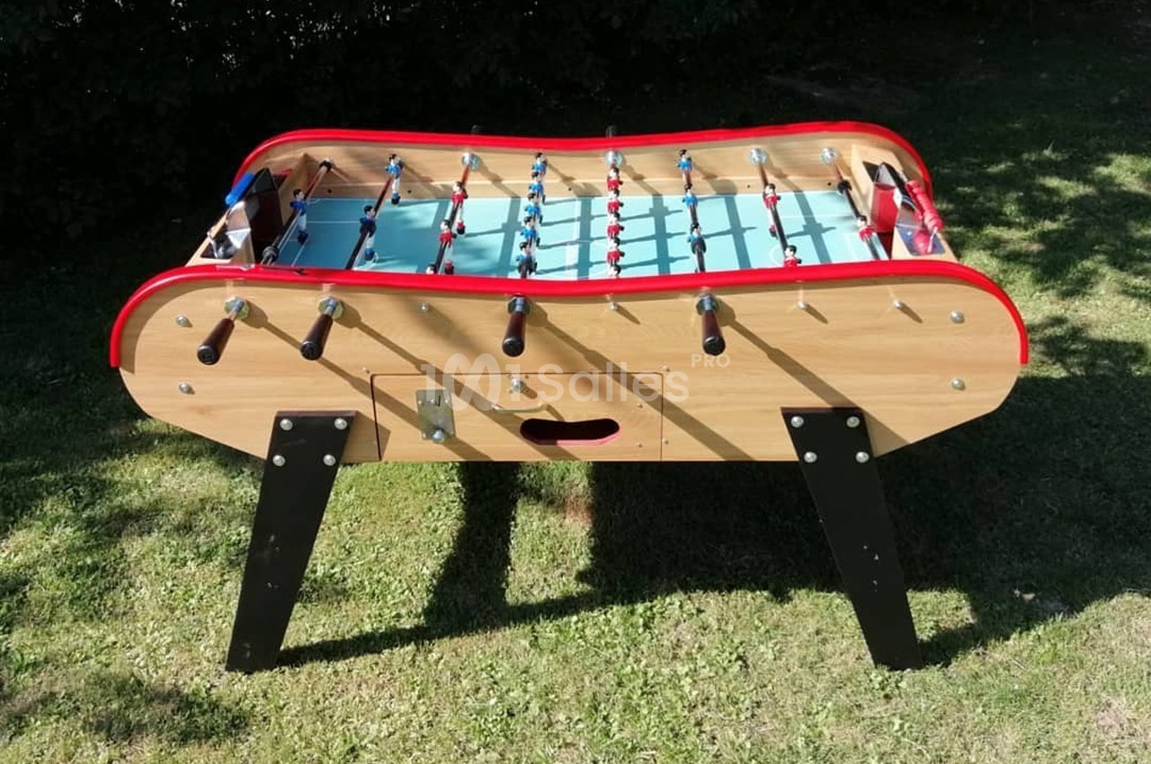 Baby-foot en bois avec surface de jeu bleue et joueurs rouges et bleus, posé sur une pelouse verte.