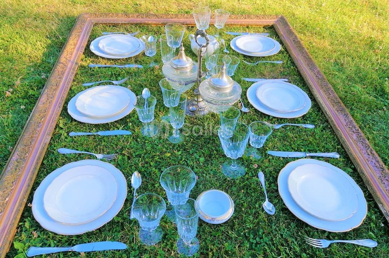 Table dressée sur de l'herbe avec vaisselle en porcelaine, couverts en métal et verres en cristal, encadrée par un cadre…