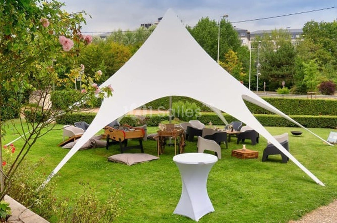 Tente blanche en étoile installée sur une pelouse, entourée de mobilier de jardin et de verdure.