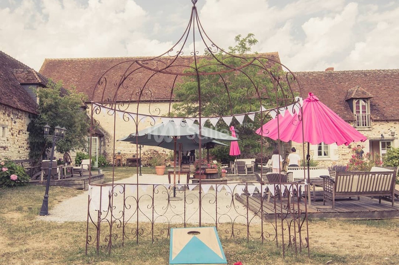Cour extérieure avec mobilier de jardin, parasols colorés et structure métallique décorative devant une maison en pierre.