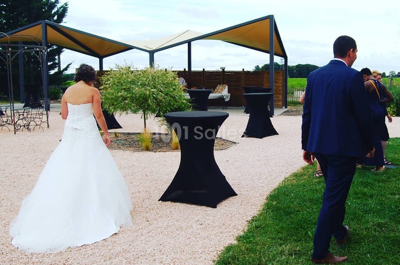 Une mariée en robe blanche et un homme en costume marchent dans un espace extérieur aménagé avec tables hautes et tonnelles.