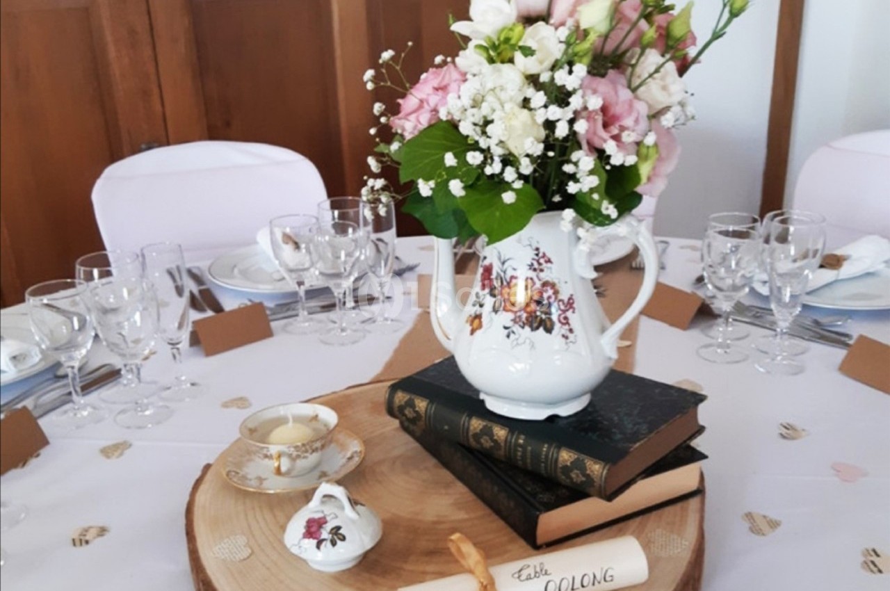 Centre de table avec bouquet de fleurs dans une théière, posé sur des livres et une rondelle de bois, entouré de vaisselle.
