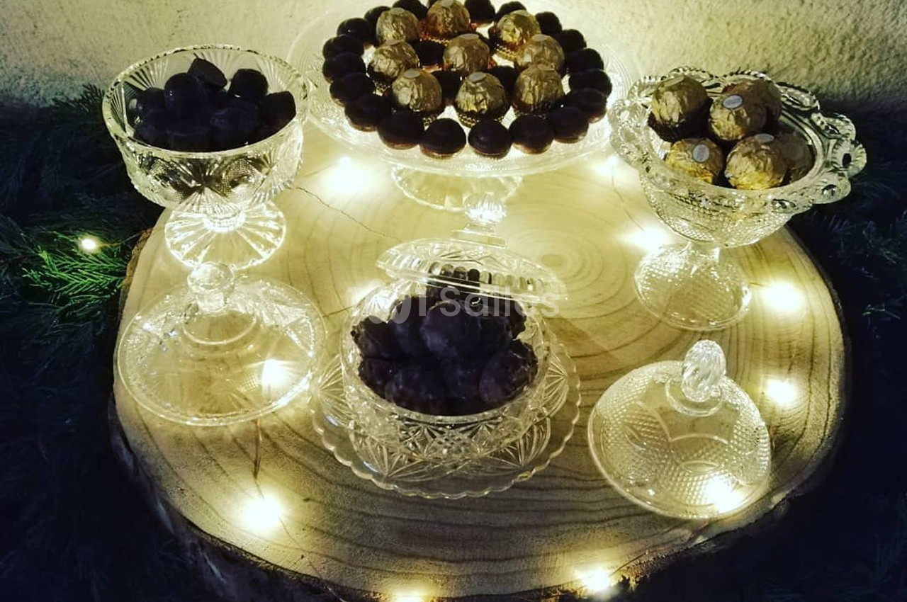 Assortiment de chocolats présentés dans des coupes en verre sur une tranche de bois éclairée par des guirlandes lumineuses.
