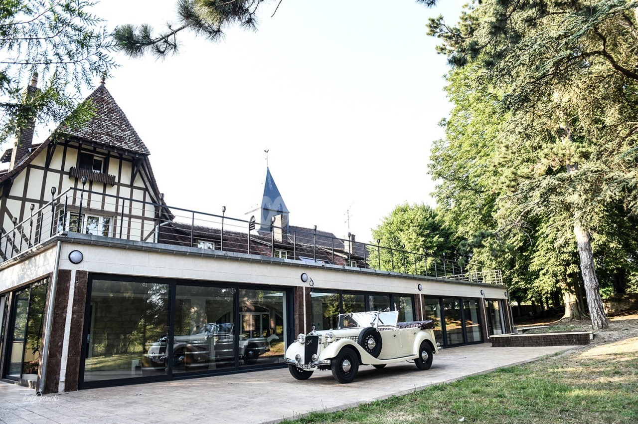 Voiture ancienne blanche garée devant un bâtiment à colombages avec une terrasse et des arbres en arrière-plan.