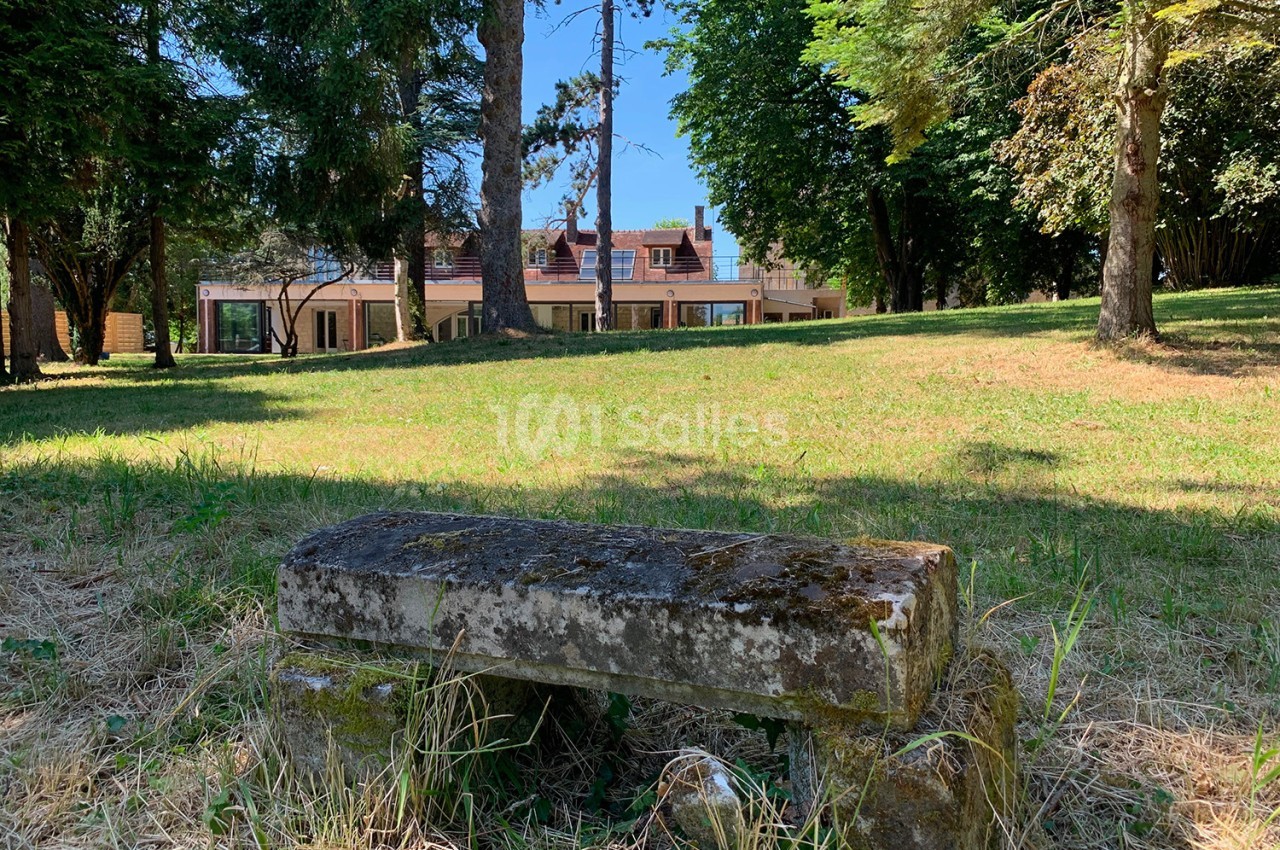 Banc en pierre ancienne dans un parc verdoyant, avec une maison moderne visible en arrière-plan.