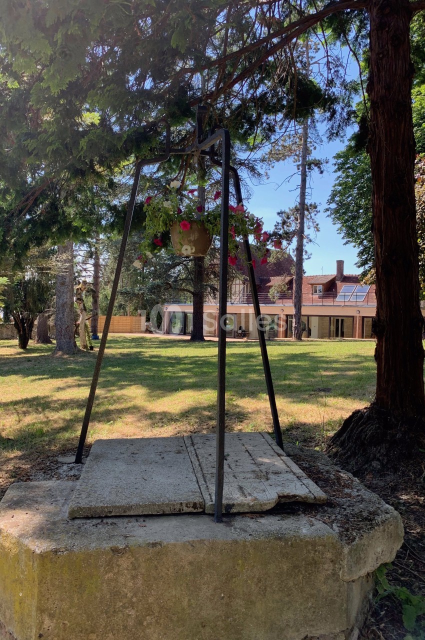 Un puits en pierre surmonté d'une structure métallique avec un pot de fleurs, entouré d'arbres dans un jardin.