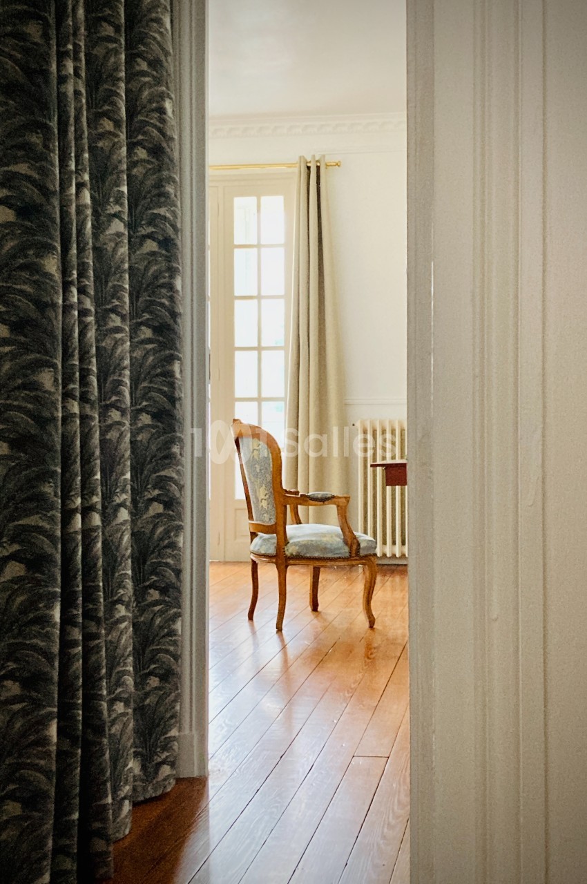 Une chaise en bois avec assise rembourrée est placée près d'une fenêtre dans une pièce lumineuse au sol en parquet.