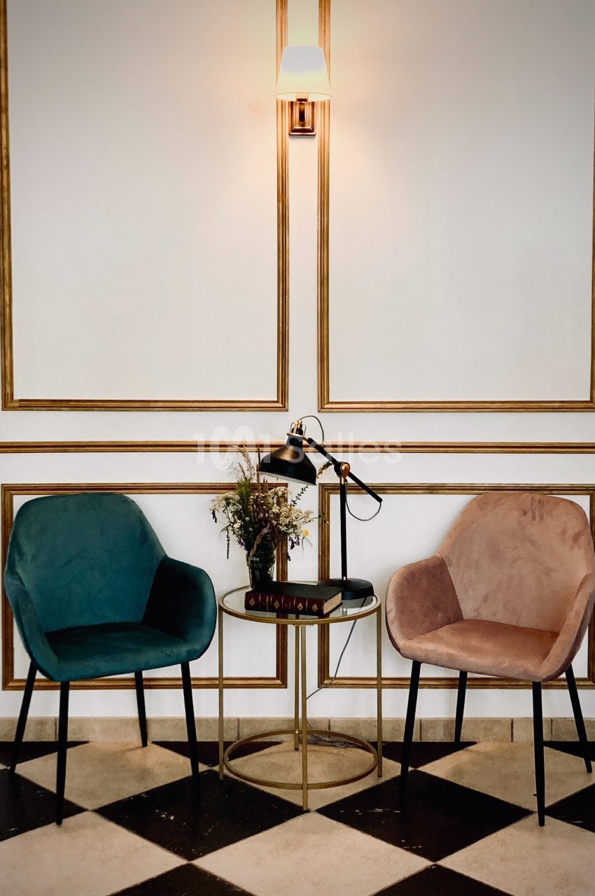 Deux fauteuils colorés entourent une table ronde avec une lampe, des livres et un bouquet, sur un sol en damier.