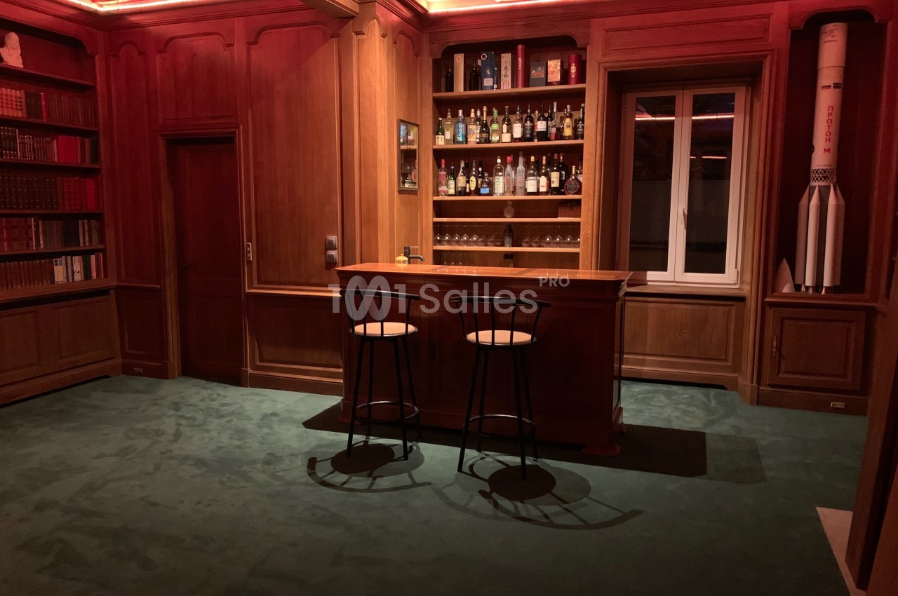 Bar en bois avec étagères remplies de bouteilles, deux tabourets et une décoration de fusée dans un intérieur chaleureux.
