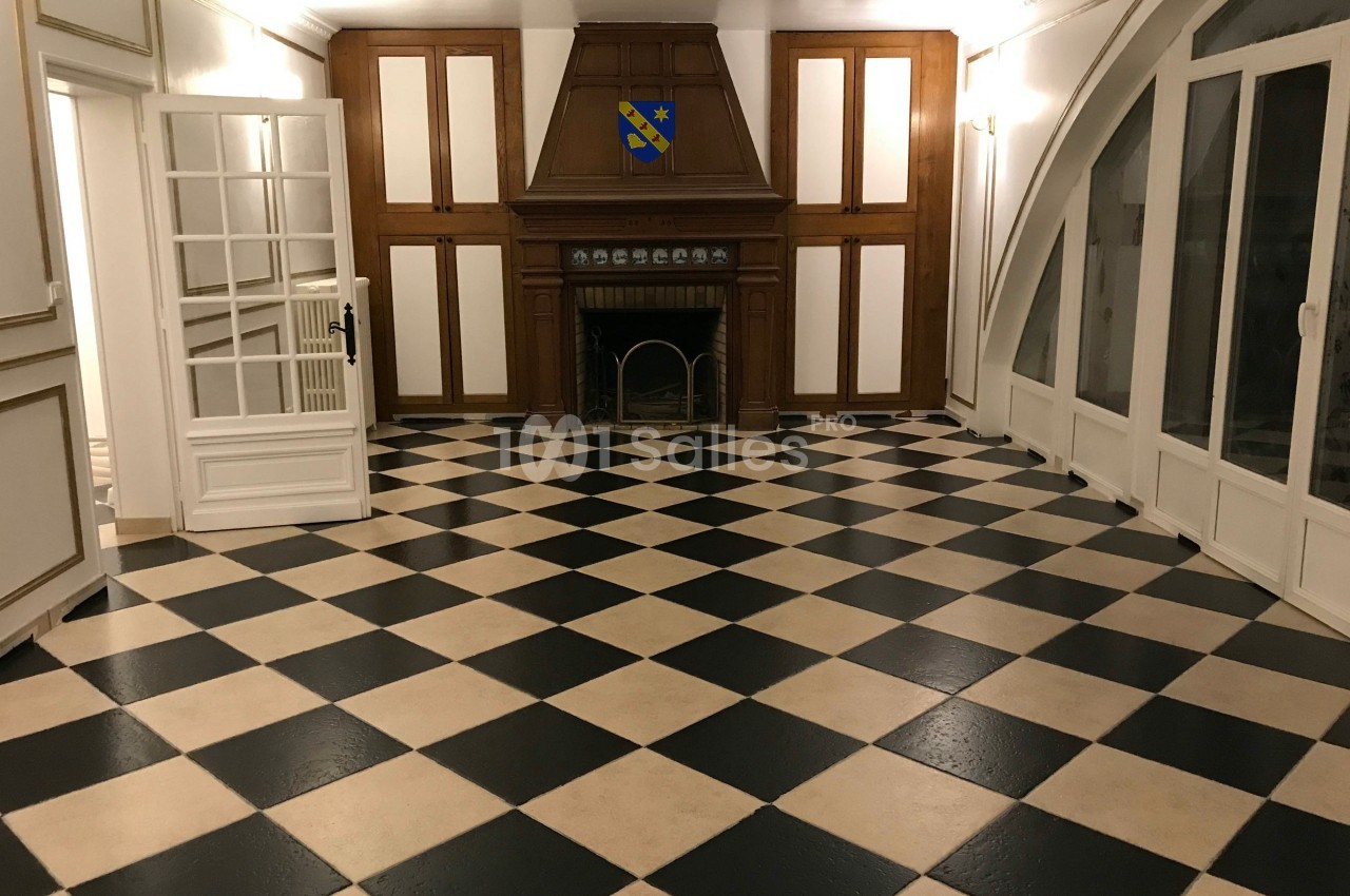 Salle avec un sol en damier noir et beige, cheminée en bois, murs blancs et grandes fenêtres cintrées.