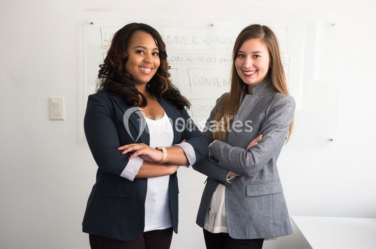 Deux femmes souriantes debout devant un tableau blanc, bras croisés, dans un environnement professionnel.