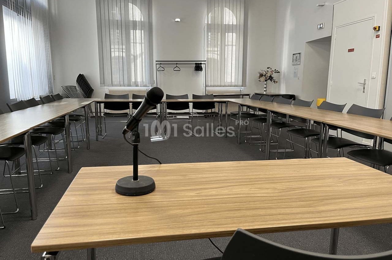 Salle de réunion lumineuse avec tables disposées en U, un micro sur une table et des chaises alignées.