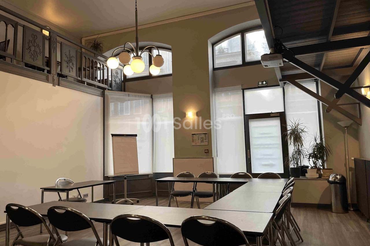 Salle de réunion lumineuse avec tables en U, chaises pliantes, tableau de conférence et grandes fenêtres.