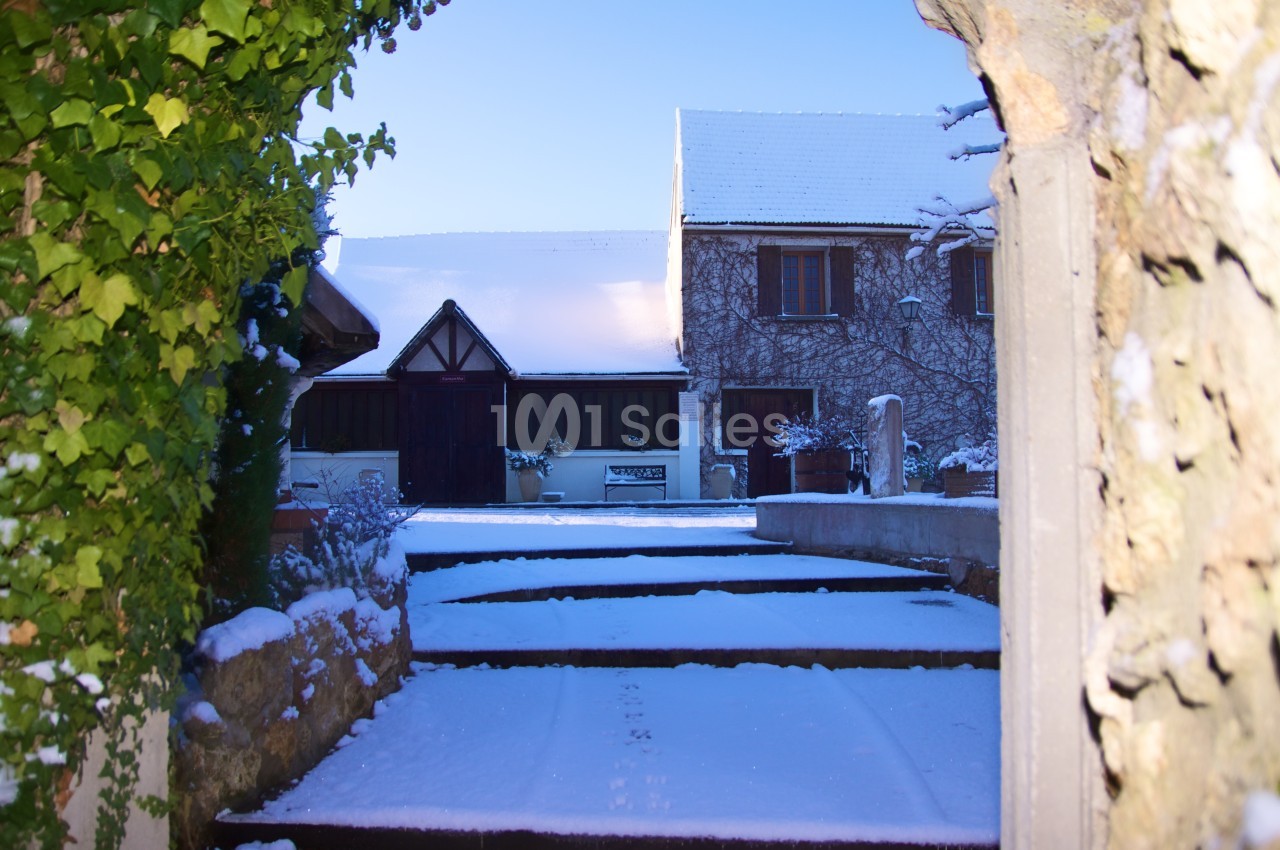Cour enneigée entourée de murs en pierre, avec une maison en arrière-plan sous un ciel bleu clair.