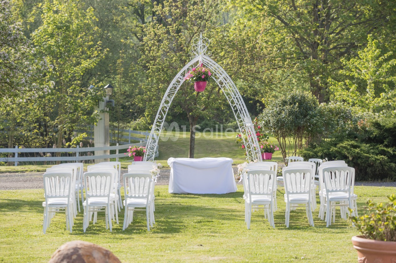 Chaises blanches disposées en rangées devant une arche décorée de fleurs dans un jardin verdoyant.