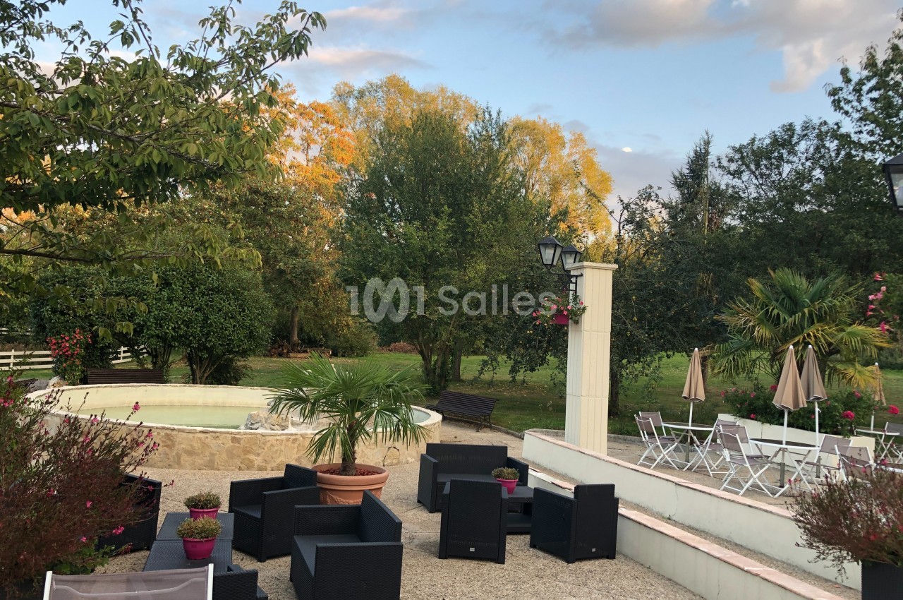Espace extérieur avec mobilier de jardin, fontaine, végétation luxuriante et ciel partiellement nuageux en arrière-plan.