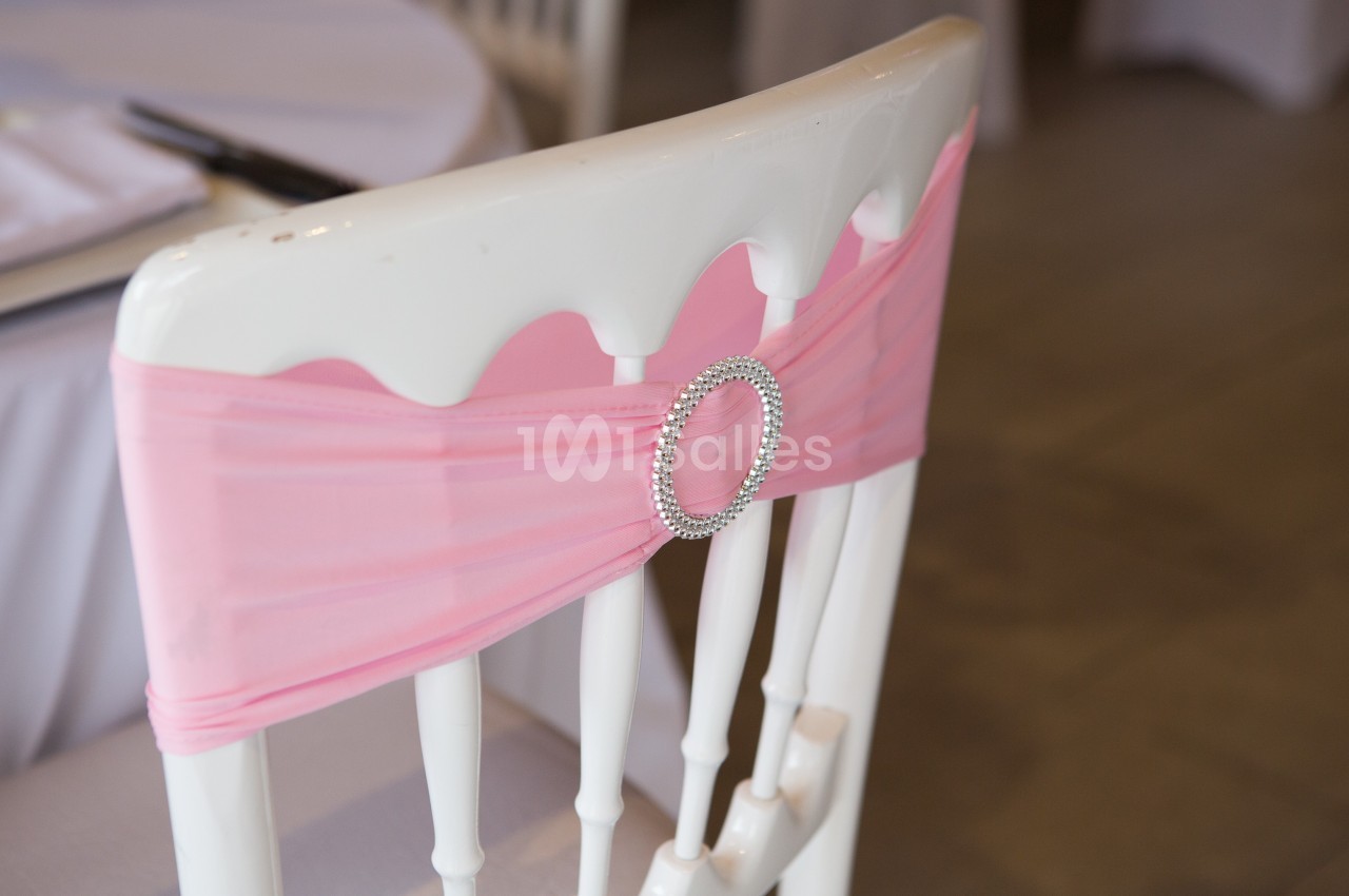 Chaise blanche décorée d'un nœud en tissu rose avec une boucle ornée de strass, dans un cadre de réception.