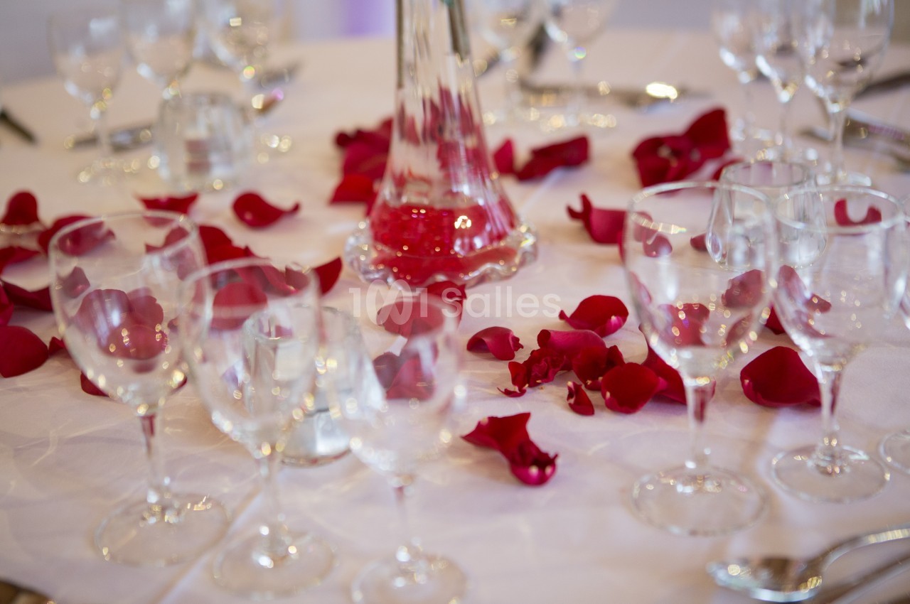 Pétales de roses rouges dispersés sur une table dressée avec des verres et un vase en verre transparent.