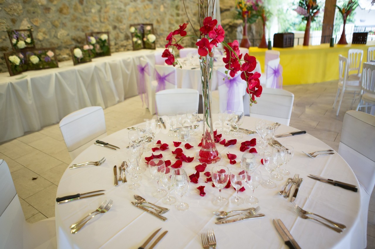 Table ronde décorée avec une nappe blanche, des pétales rouges, un vase central fleuri et des couverts disposés.