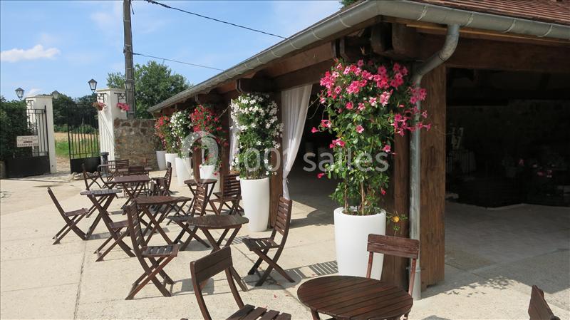 Terrasse extérieure avec tables et chaises en bois, décorée de grandes jardinières fleuries sous un auvent.