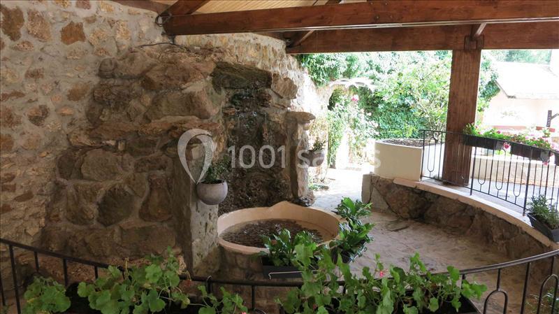 Mur en pierre avec une petite fontaine entourée de plantes, sous un abri en bois ouvert sur un jardin.