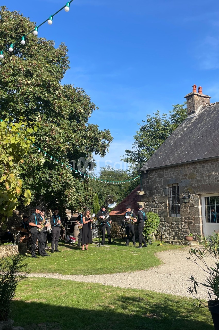 Un groupe de musiciens joue en plein air dans une cour verdoyante près d'une maison en pierre sous un ciel bleu.