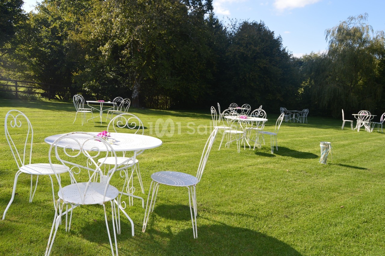 Tables et chaises blanches en métal disposées sur une pelouse verdoyante, entourées d'arbres.