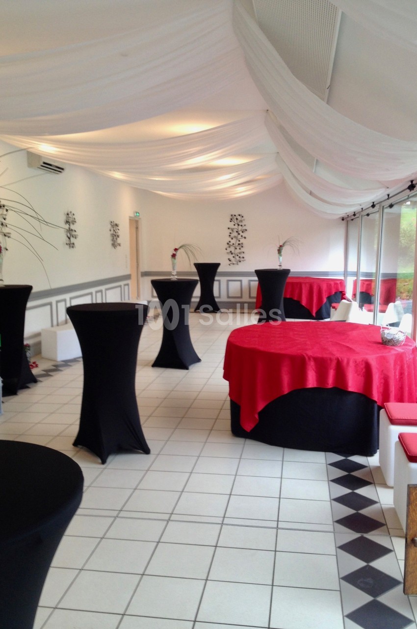Salle de réception lumineuse avec tables hautes noires et tables rondes recouvertes de nappes rouges.