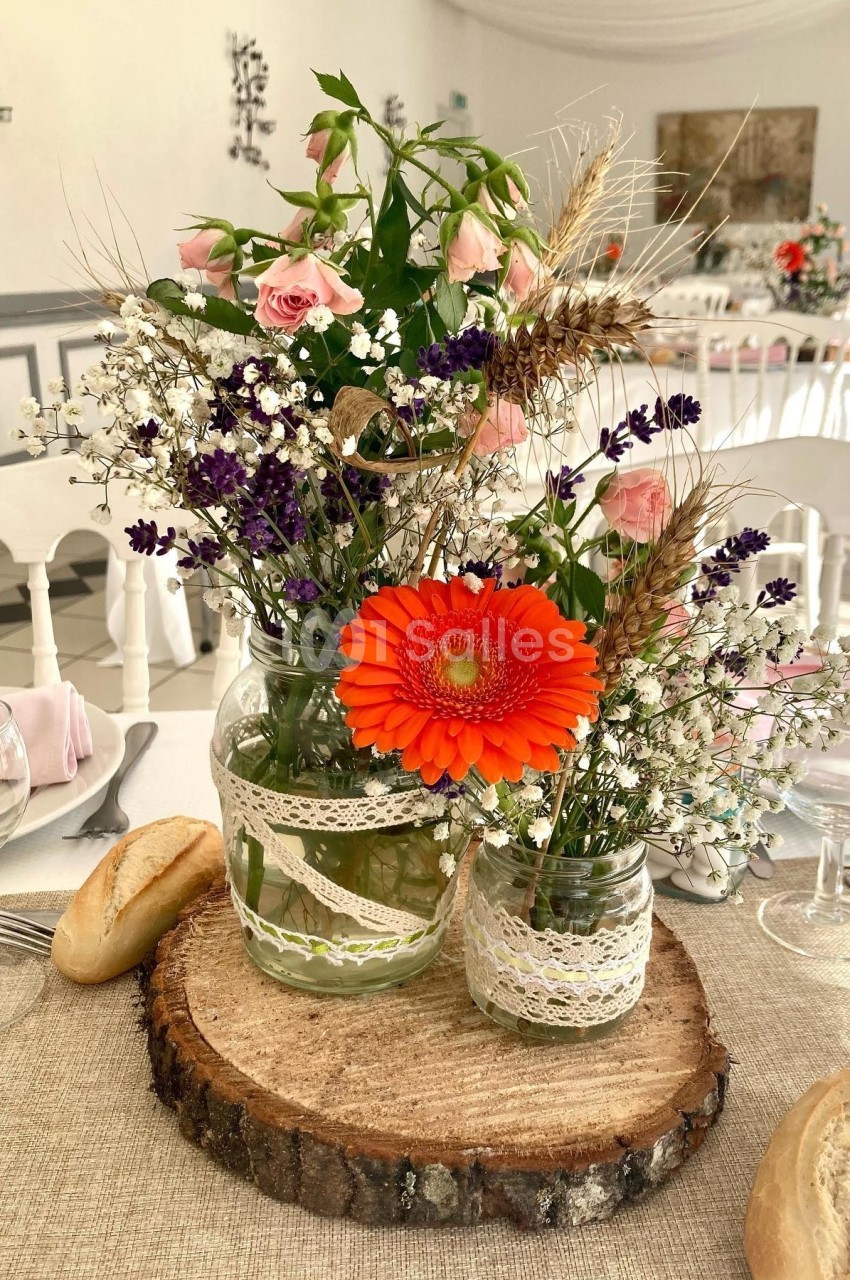 Centre de table champêtre avec fleurs colorées dans des bocaux en verre décorés, posé sur une rondelle de bois.