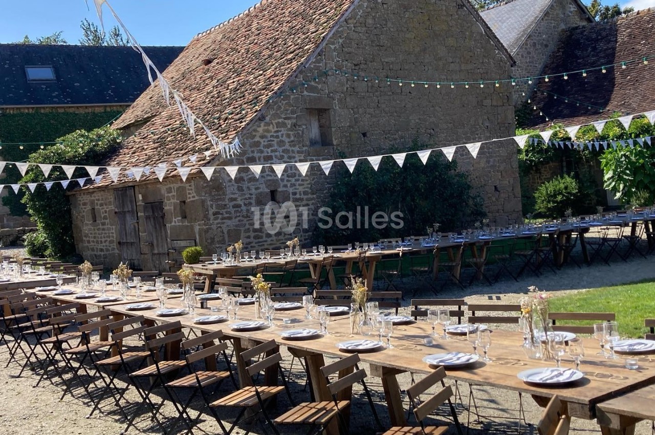 Tables en bois dressées avec vaisselle et décorations, alignées en extérieur devant une bâtisse en pierre sous des…