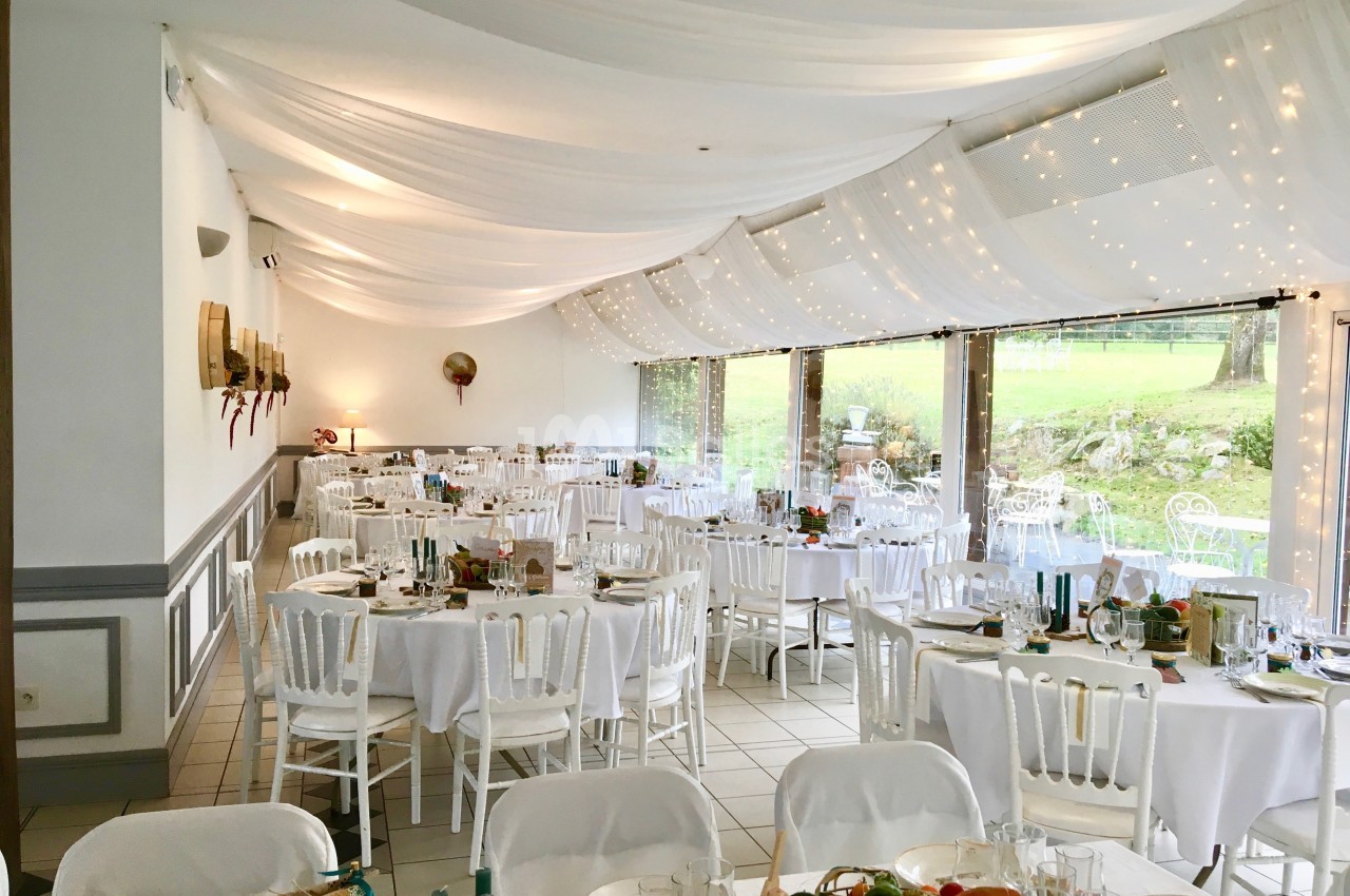 Salle de réception décorée avec des tables rondes dressées, des chaises blanches et un plafond drapé de voilages lumineux.