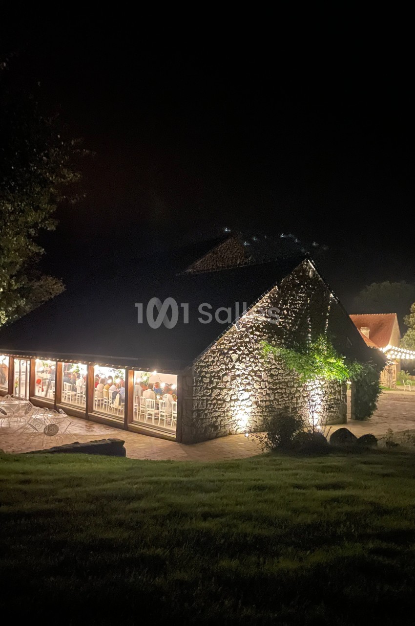 Bâtiment en pierre éclairé la nuit, avec des fenêtres révélant une salle illuminée et des invités à l'intérieur.