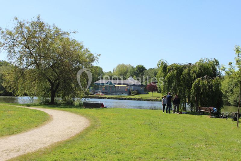 Un sentier longe un parc verdoyant près d'un lac, avec des arbres, un bâtiment en arrière-plan et quelques personnes debout.