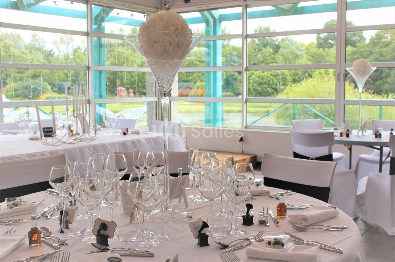 Salle de réception lumineuse avec tables dressées, nappes blanches, verres, et centre de table floral en hauteur.